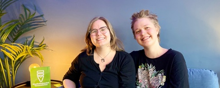 Två leende personer sitter tillsammans i en blå soffa i ett rum med blå vägg och grön växt.