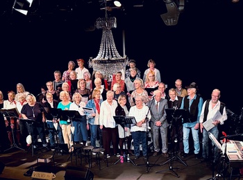 Kör och musiker uppträder på scen inför publik i en teaterlokal med mikrofoner och instrument.