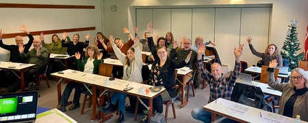 En grupp människor sitter i ett klassrum och vinkar mot kameran, med papper och datorer på borden.