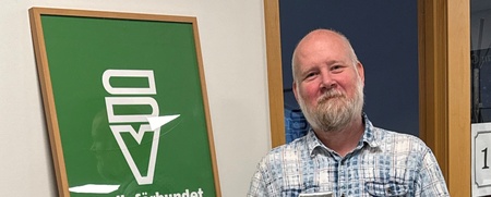 Man står leende med ett paket i handen bredvid en skylt för Studieförbundet Vuxenskolan.