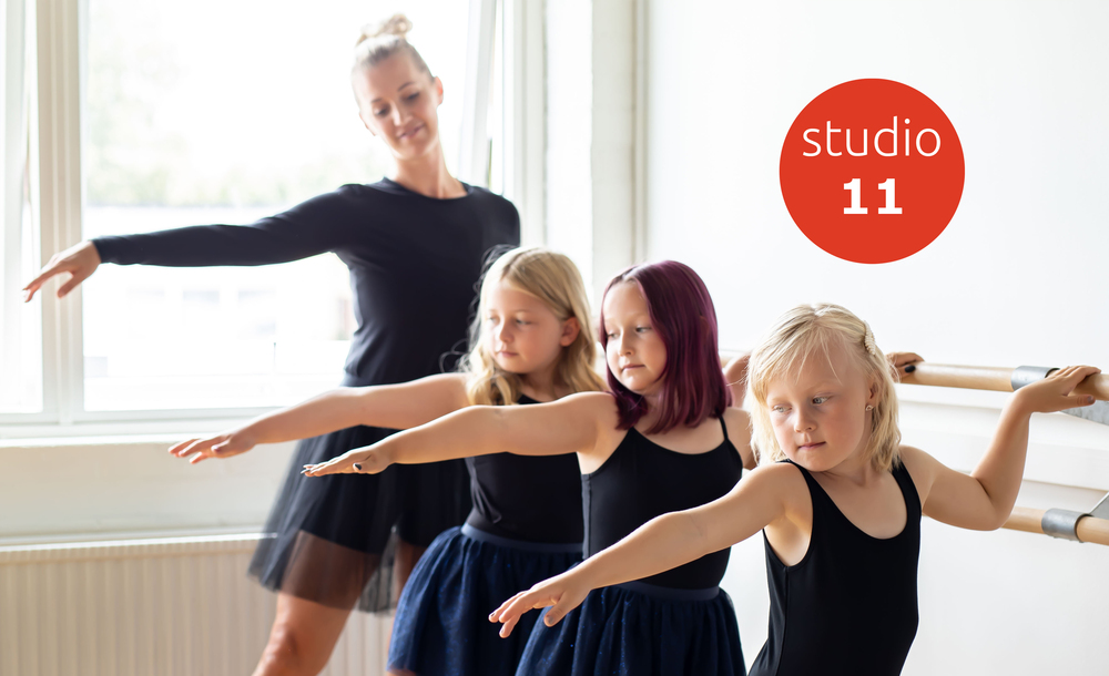 Tre barn och en vuxen övar balett vid en balettstång i en ljus dansstudio med skylten 'studio 11' på väggen.