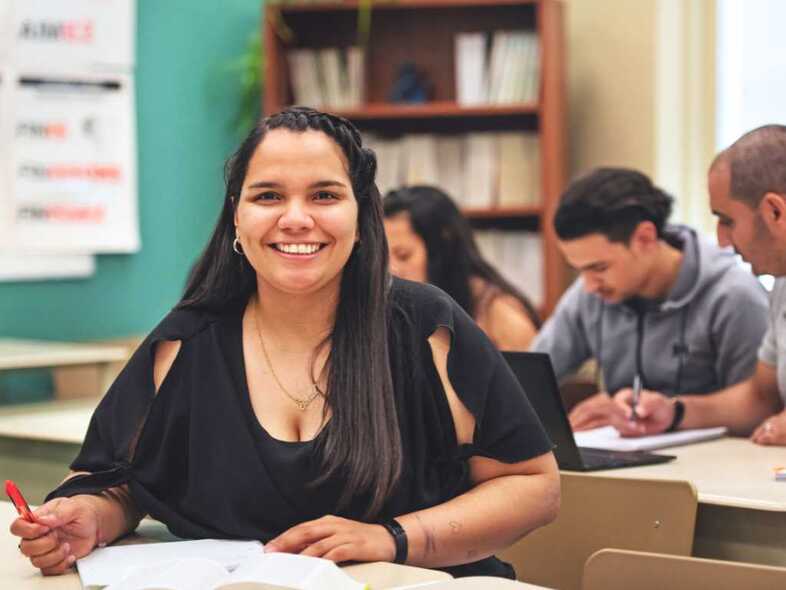 En leende person sitter i ett klassrum och arbetar. I bakgrunden är det fler personer som sitter och arbetar.