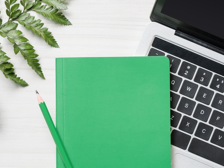 Grön anteckningsbok och penna på en bärbar dator, med växt och ormbunksblad på ett vitt skrivbord.