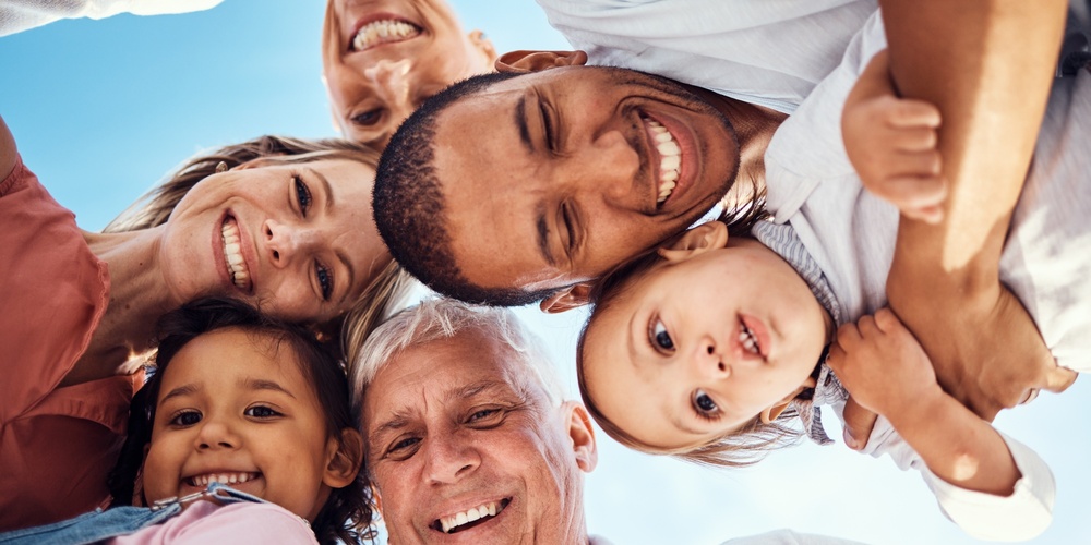 Sex personer i olika åldrar står i en cirkel och ler mot kameran under en blå himmel.