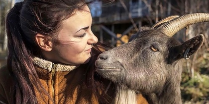 En kvinna i brun fleecejacka tittar leende på en get som står nära henne utomhus.