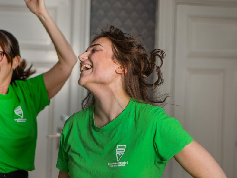 Två glada kvinnor i gröna t-shirts skrattar och dansar inomhus framför vita dörrar.