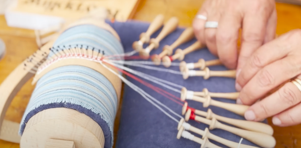 Närbild på händer som tillverkar spets med hjälp av knyppelpinnar och en rund kudde.