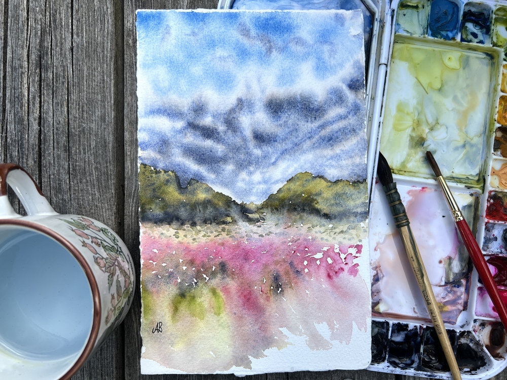 Akvarellmålning av landskap med blå himmel, gröna kullar och rosa fält, omgiven av penslar, färgpalett och en kopp.