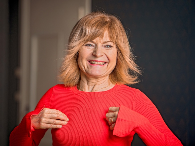Leende kvinna med axellångt blont hår i röd tröja står inomhus och ser glad ut.