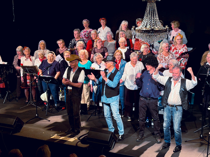 En stor kör står på en scen under en kristallkrona, några personer står framför och applåderar.