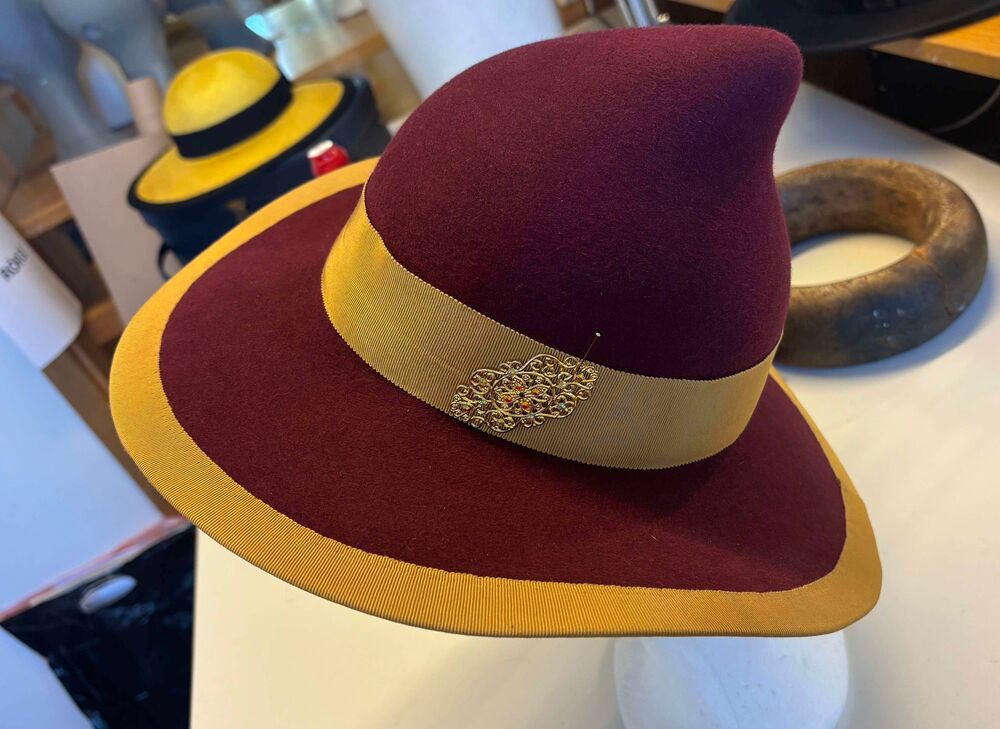 Vinröd filthatt med bred gul kant och band, dekorerad med ett guldfärgat emblem, står på ett bord.