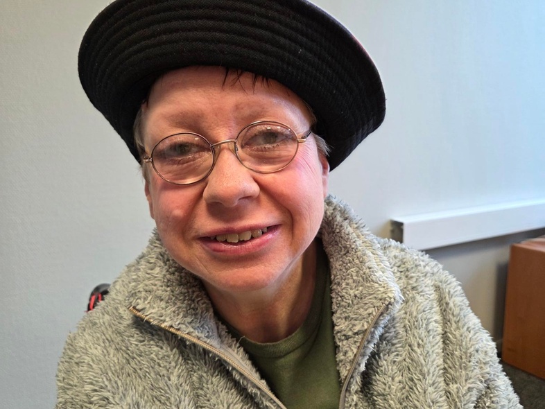 Person med grå fluffig jacka och svart hatt sitter inomhus, ansiktet är suddat.