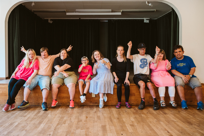 En grupp unga personer sitter på en scenkant och poserar glatt tillsammans inomhus.