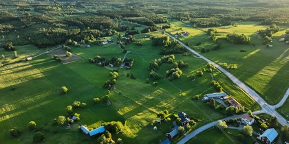 Flygfoto över ett grönt landskap med åkrar, skog och utspridda hus i kvällssol.