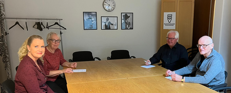 Fyra äldre personer sitter runt ett konferensbord i ett mötesrum med ljusa väggar och tavlor.