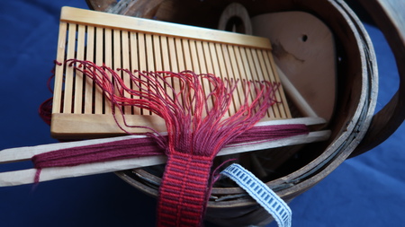 Närbild av ett bandvävningsredskap med rött garn och trädetaljer på blå bakgrund.