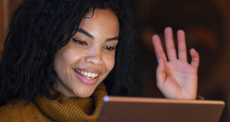 Kvinna med lockigt hår vinkar och ler mot en surfplatta, bär en gul stickad tröja.