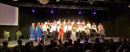 En kör står på scen och sjunger inför en sittande publik i en mörk konsertsal.