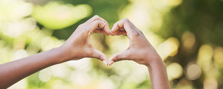 Två händer formar ett hjärta mot en suddig, grön bakgrund utomhus.
