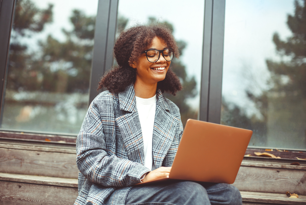 Leende person med glasögon sitter utomhus på en trappa och arbetar på en bärbar dator.