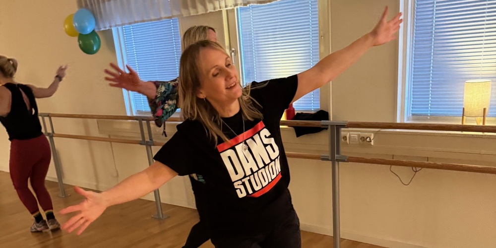Kvinna dansar glatt i en dansstudio med trägolv och speglar, iförd svart t-shirt med texten 'DANS STUDIO'.