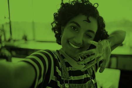 Person med lockigt hår tar en selfie, ler och visar en hand med färg på, i en inomhusmiljö med grönt filter.