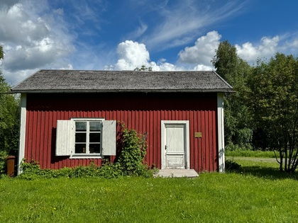 Röd liten stuga med vita knutar, vita fönsterluckor och dörr, omgiven av grön gräsmatta och träd under blå himmel.