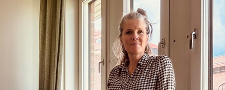 Person i svartvit mönstrad skjorta står vid ett fönster i ett ljust rum med gröna gardiner och papperspåsar.