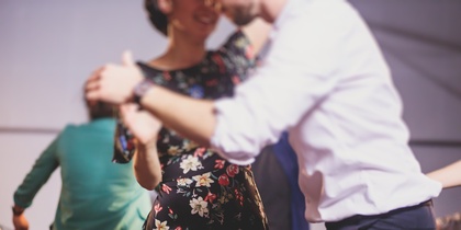 Två personer dansar tillsammans, en i blommig klänning och en i vit skjorta, i en suddig inomhusmiljö.