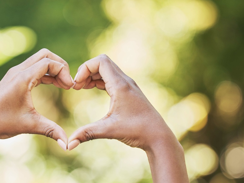Två händer formar ett hjärta mot en suddig, grön bakgrund utomhus.