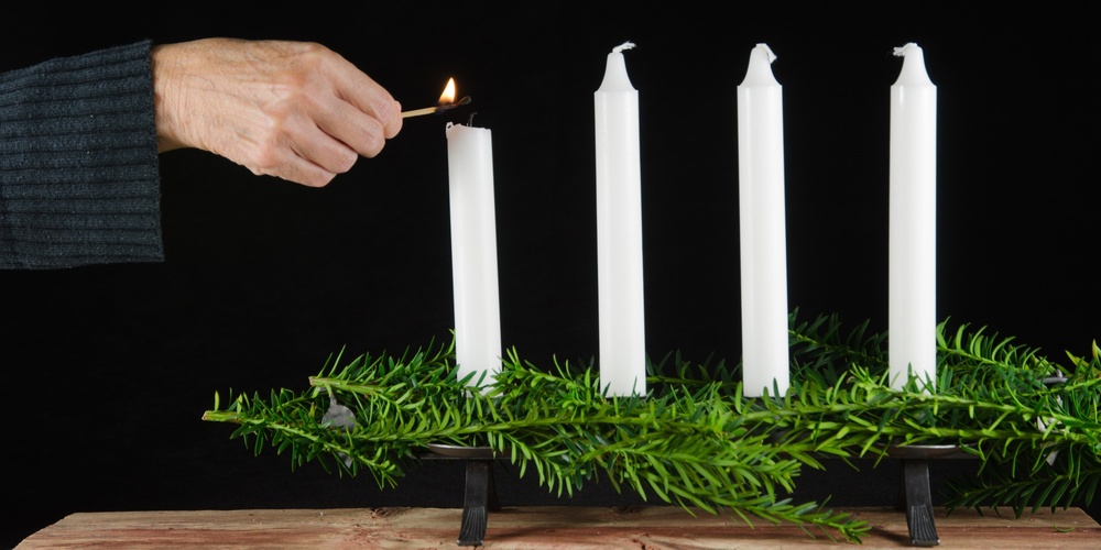 En hand tänder det första av fyra vita ljus i en adventsljusstake med gröna kvistar.