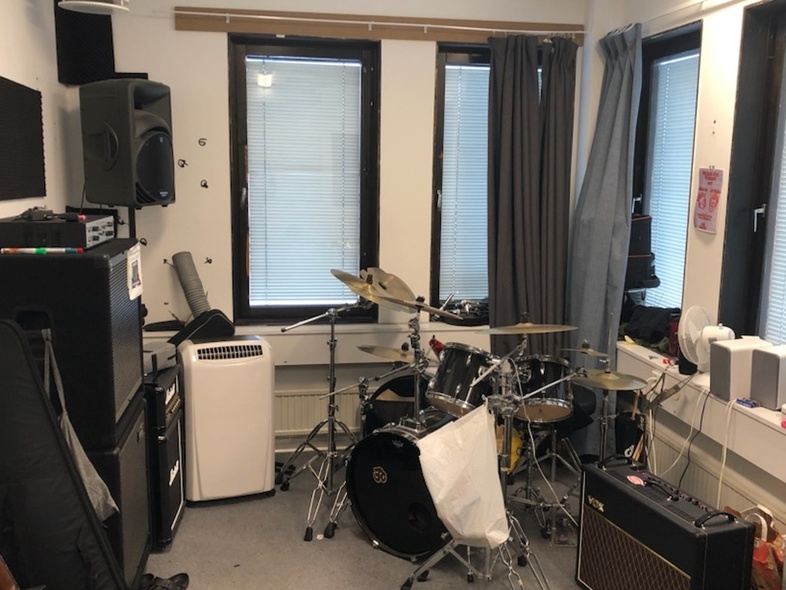 Ett musikrum med trumset, gitarrförstärkare, högtalare och ljudutrustning, samt fönster med gardiner.