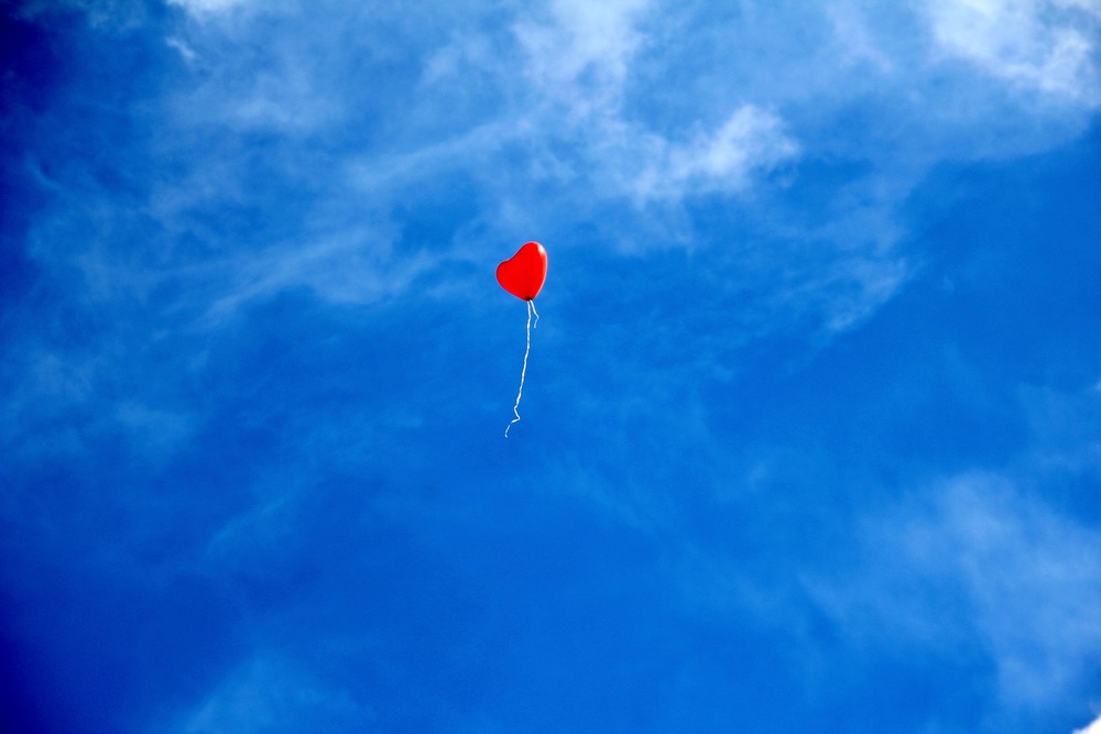 En röd hjärtformad ballong svävar ensam mot en klarblå himmel med några moln.