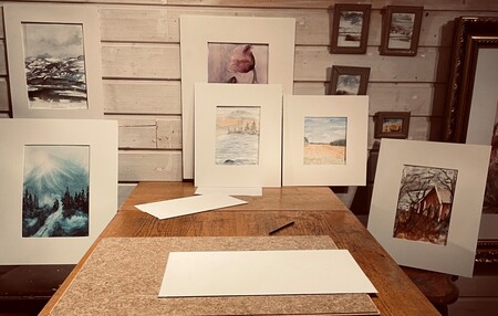 Ett bord med flera inramade akvarellmålningar uppställda mot en trävägg, samt papper och penna på bordet.