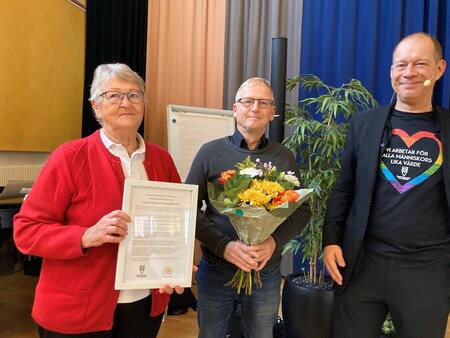 Tre personer står inomhus, en håller ett diplom och en annan en bukett blommor.