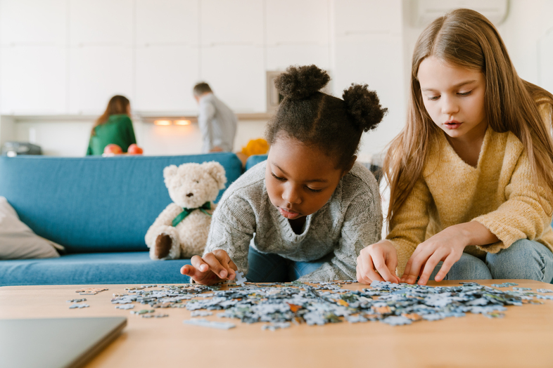 Två barn lägger pussel på ett bord i ett vardagsrum, med en nallebjörn i bakgrunden.