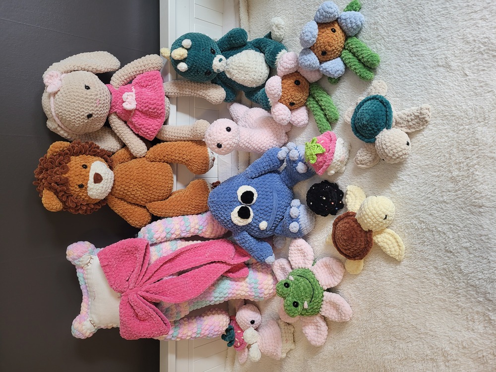 Flera virkade mjukdjur, inklusive en björn, kanin, dinosaurie, groda, sköldpaddor och blommor, placerade på en ljus matta.