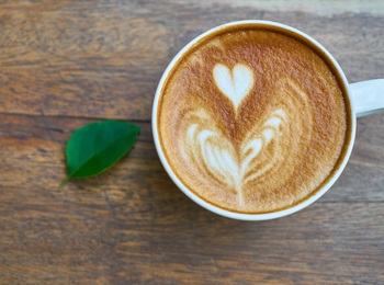 En kopp cappuccino med hjärtformad latte art står på ett träbord bredvid ett grönt blad.