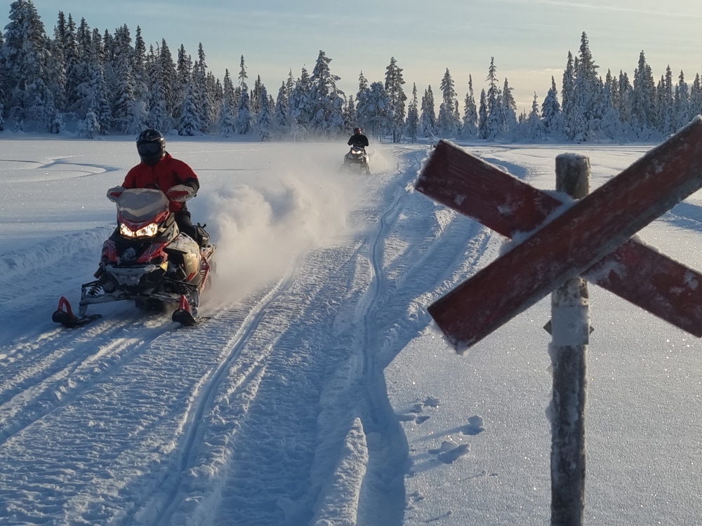Två personer kör snöskoter på ett snötäckt fält med skog i bakgrunden och en röd krysskylt i förgrunden.