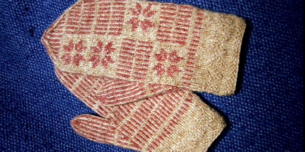 Ett par stickade vantar med röd och beige mönster ligger på ett blått tygunderlag.