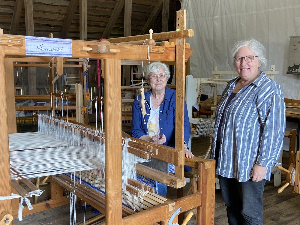 Två personer står vid en stor vävstol i en träbyggnad, omgivna av vävutrustning.