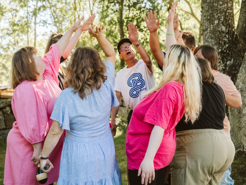 En grupp människor står i en ring utomhus och sträcker upp händerna mot mitten under ett träd.