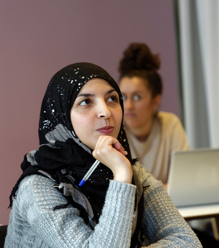 Ung kvinna med hijab sitter i klassrum och tänker, håller en penna mot hakan.