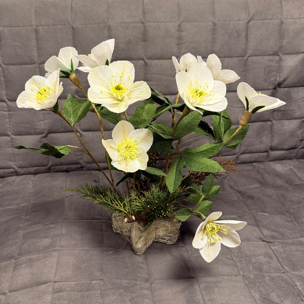 En bukett vita konstgjorda blommor med gröna blad i en dekorativ bas, placerad mot en grå vadderad bakgrund.