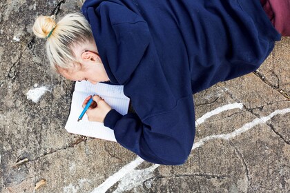 Person med blont hår i knut ligger på marken och skriver i en anteckningsbok med blå penna.