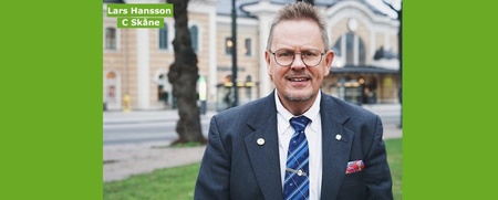 Man i kostym står utomhus framför en byggnad, ler mot kameran. Grön bakgrund med text: Lars Hansson, C Skåne.