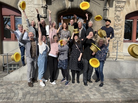 En grupp glada människor kastar gula hattar i luften utanför en stenbyggnad.