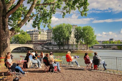 Folk sitter på uteservering vid floden med utsikt över bro, träd och klassiska byggnader i solsken.