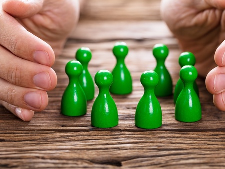 Två händer skyddar en grupp gröna spelpjäser på ett träbord.