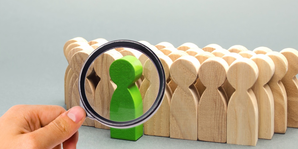 En hand håller ett förstoringsglas över en grön träfigur bland flera beige träfigurer.